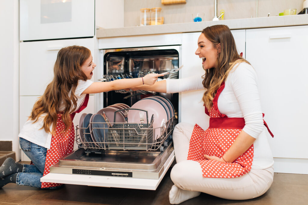 Happy pregnant mother and daughter in red aprons placing dishes in dishwasher at kitchen. Cleaner supporting NDIS participant in Perth